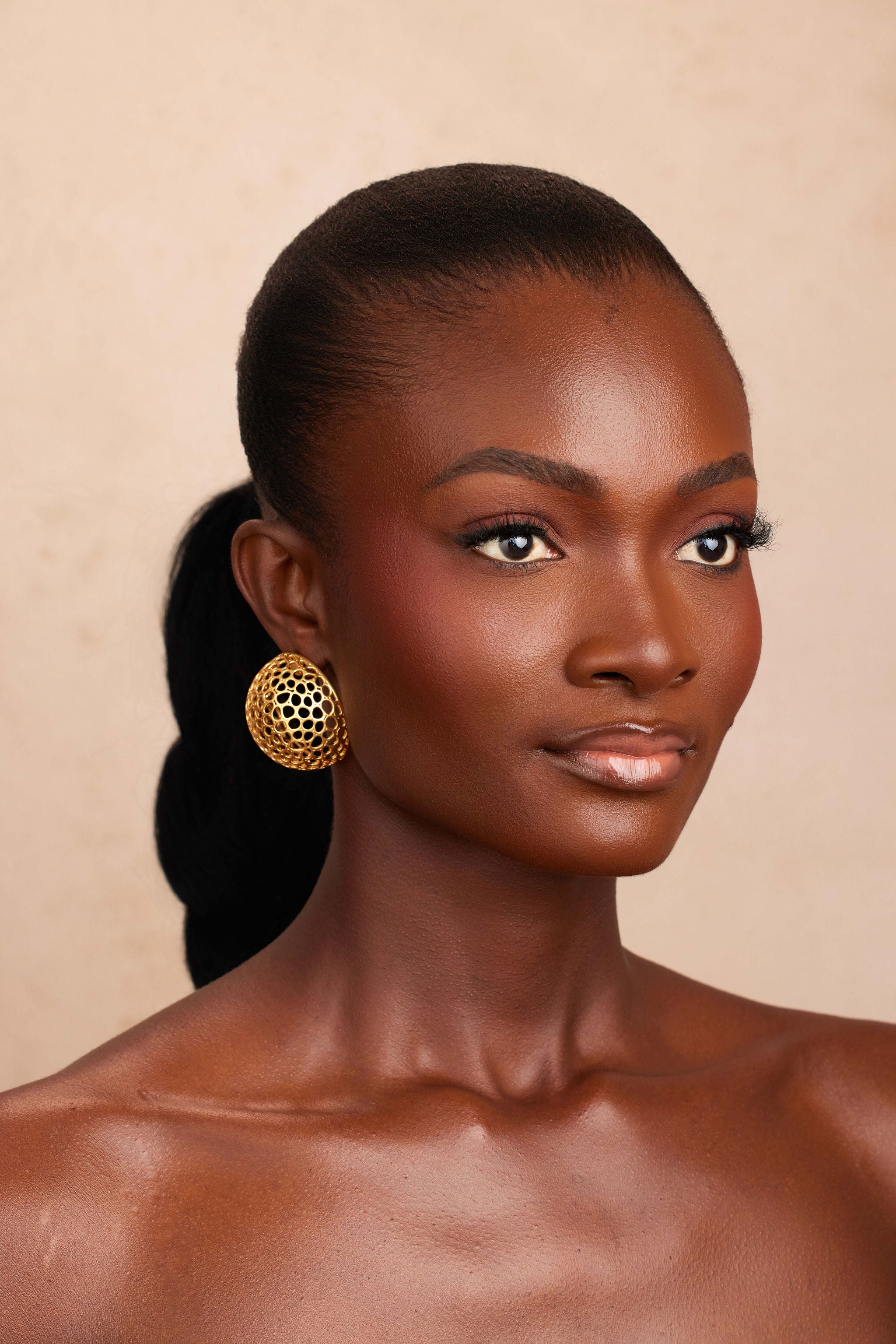 Woman with a ponytail wearing gold earrings against a beige background
